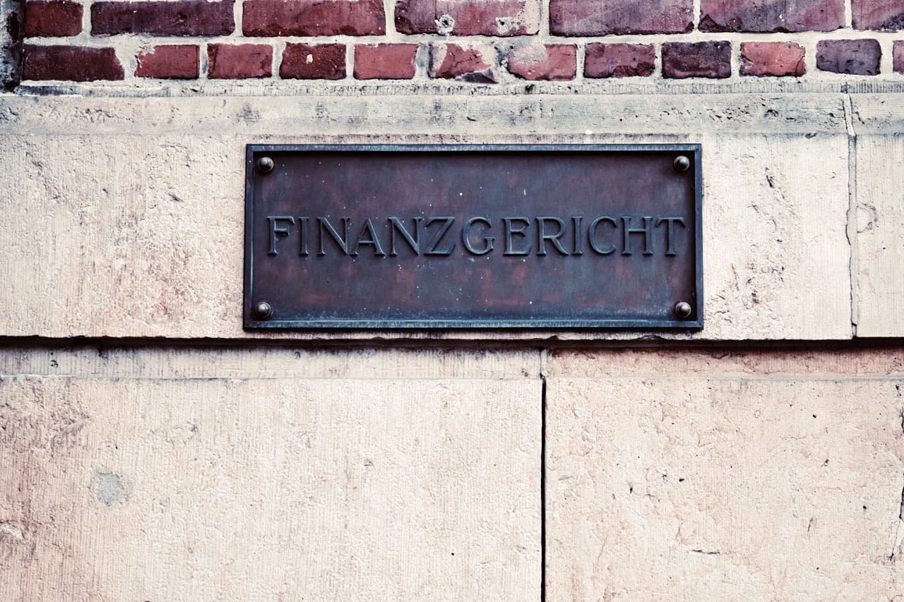 Verwitterte Bronzetafel mit der Aufschrift „Finanzgericht“ an einer Sandstein- und Ziegelfassade.